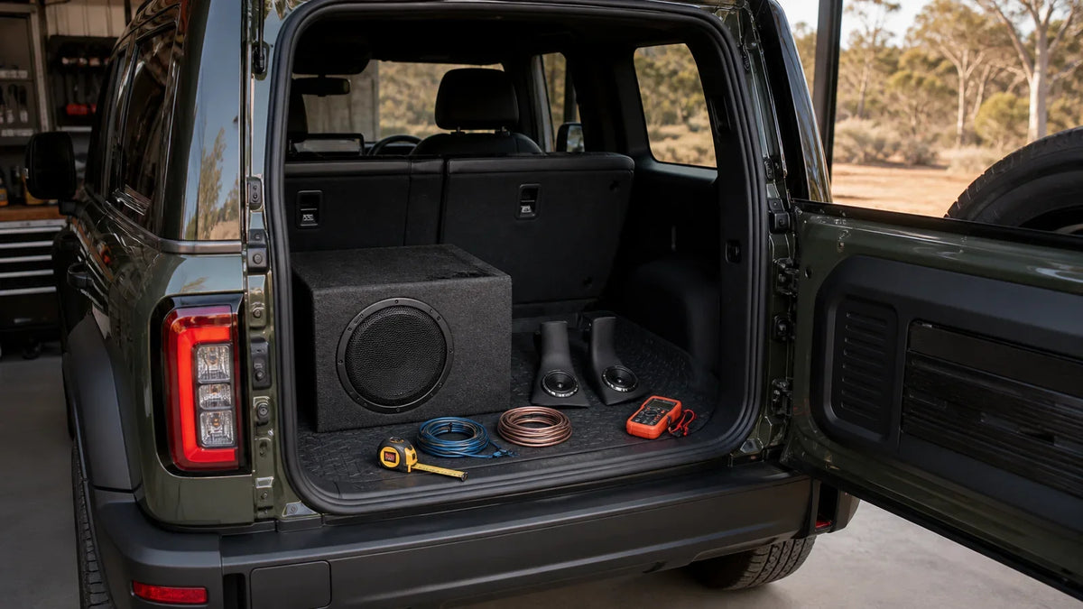 Illustrative cargo-area audio upgrade planning scene with a compact subwoofer, A-pillar speakers, measuring tools and wiring in a generic off-road SUV