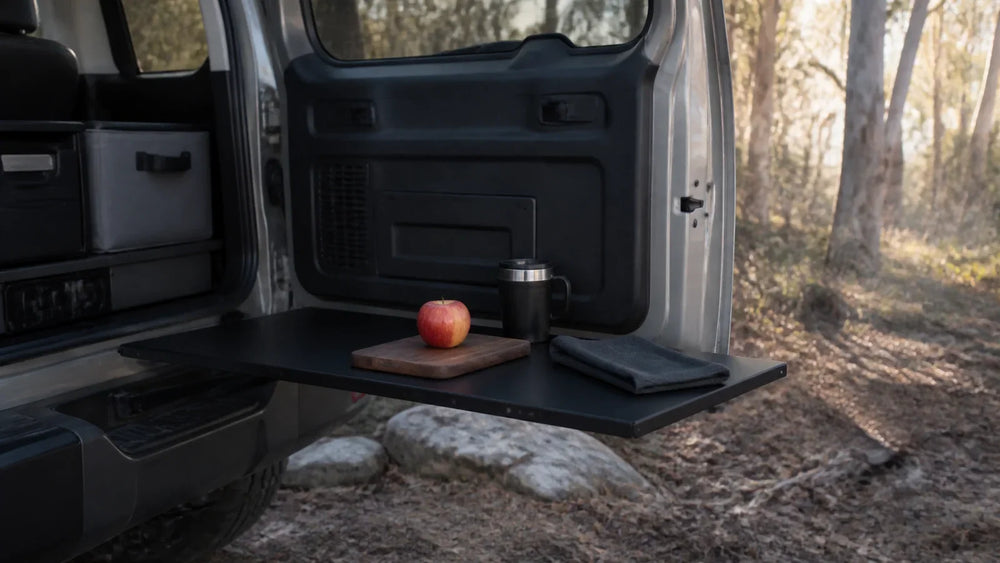 Conceptual campsite food-preparation surface on an open rear door, with an apple and covered cup